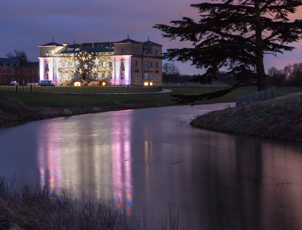 A large stately home, Croome Court, is illuminated with pink and purple light. The light reflects onto the lake in front of the house. The sky is dark.