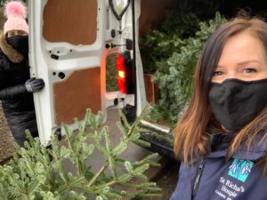 Two women stand by the back doors of a white van, holding a dark green Christmas tree. They are loading trees into the van, as part of the St Richard's Hospice Christmas Tree Collection campaign. They both wear black face masks and warm clothing.