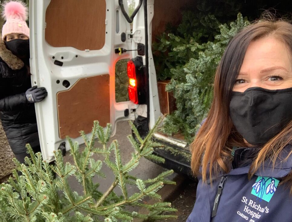 Two women stand by the back doors of a white van, holding a dark green Christmas tree. They are loading trees into the van, as part of the St Richard's Hospice Christmas Tree Collection campaign. They both wear black face masks and warm clothing.