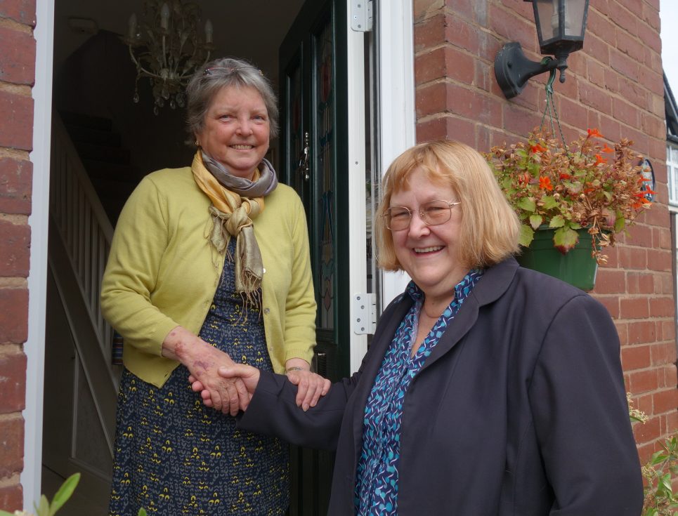 A person stands in their front doorway with a hospice Clinical Nurse Specialist. The nurse is wearing a navy blazer and blue patterned nurses' shirt. The other person is standing their doorway wearing a dress and pale yellow cardigan. They are both smiling and warmly shaking hands.