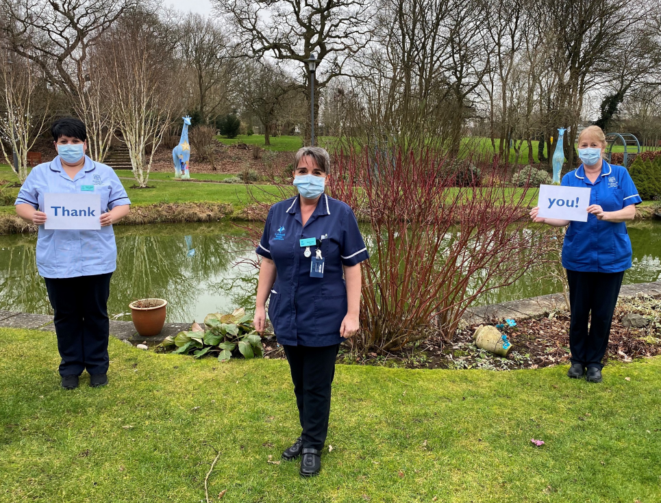 Three healthcare professionals in uniform stand together in the gardens of St Richard's Hospice. They are holding a sign which reads thank you.