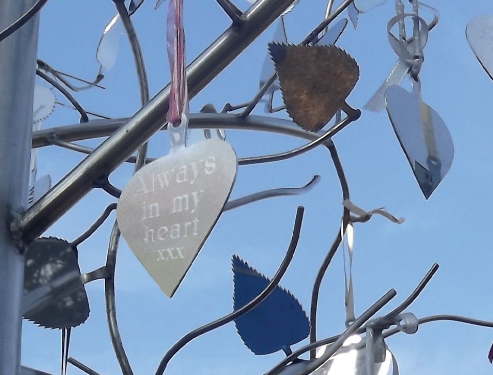 Silver metal hearts hang from the metal donor tree in the hospice gardens. The sky behind the tree is blue.
