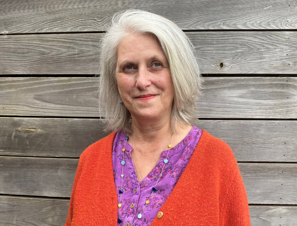 A woman with shoulder length white hair stands in front of a grey, wooden wall. She is wearing a bright red cardigan and purple top.