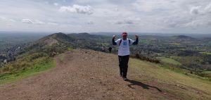 A man wearing walking gear and a white t-shirt bearing the St Richard's Hospice logo walks up a hill. He is on the Malvern Hills and there is a spectacular view of the ridge extending behind him. The sky is blue.