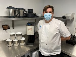 Head of Catering, Kevin Ratcliffe, stands in a chrome kitchen area next to an ice cream maker. He is wearing a white chef's top and blue face mask.