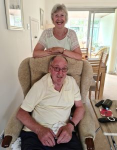 A husband and wife pictured together in a sitting room. The husband sits in an arm chair, and his wife stands behind him. They both wear light-coloured clothing, and the sitting room is decorated with light colours.