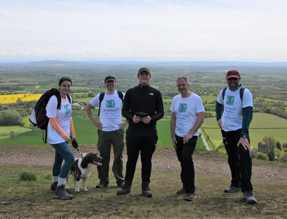 A group of people stand together wearing walking gear and white t-shirts with the St Richard's Hospice logo. They are on the Malvern Hills, with a great view of Worcestershire behind them. The sky is blue.