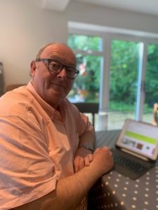 A man wearing a light coloured shirt sits at a table with a laptop computer in front of him. 