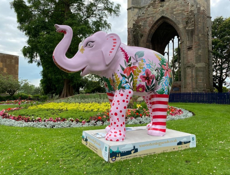 A pink, patterned elephant stands in a garden in front of a bed of flowers and St Andrew's Spire, Worcester, in the background.