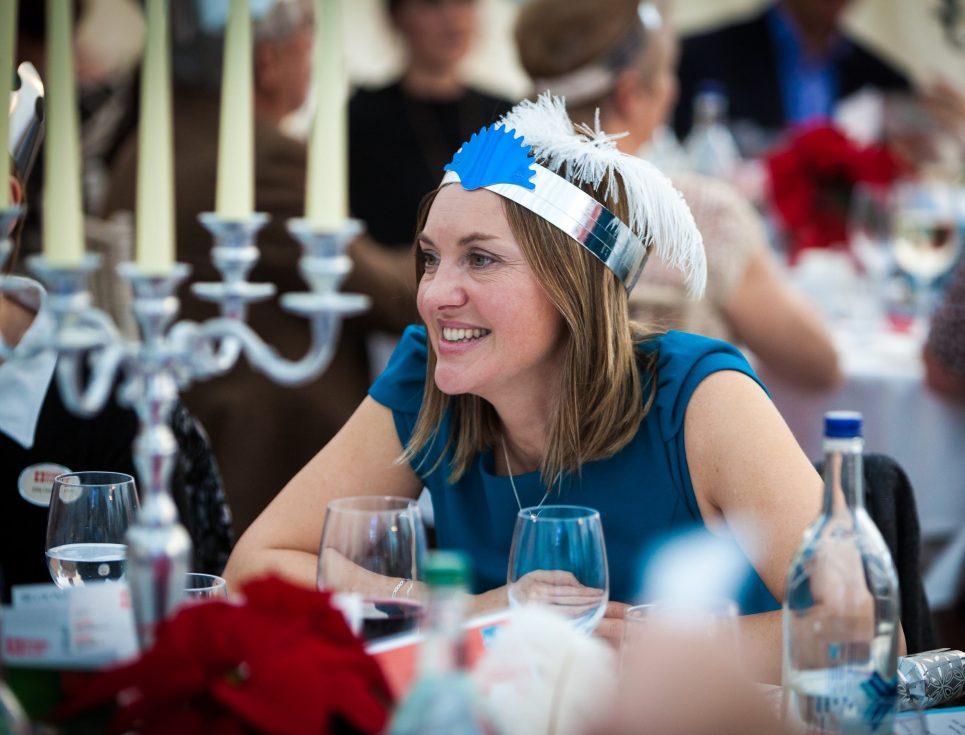 A woman wearing a blue dress and blue party hat with a white feather sits smiling at a table. There is a large, silver candlestick on the table with three candles.