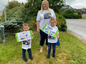 Two children, and a woman, all wearing white t-shirts stand together on a green patch of grass. The children are holding hand-drawn signs which read 'well done daddy' and 'you are amazing'.