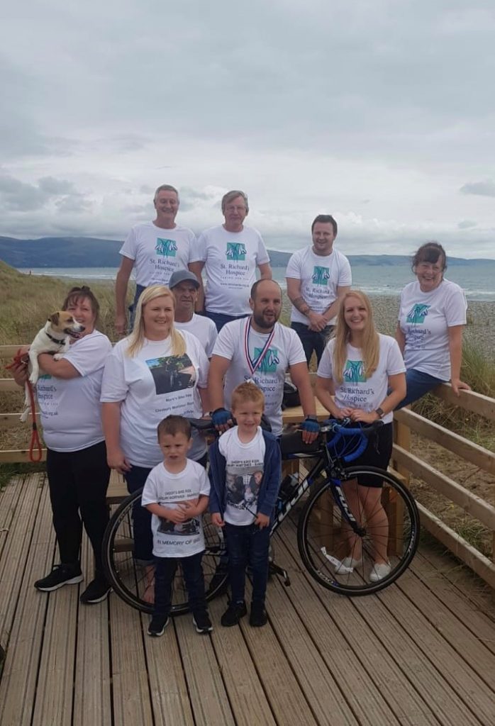 A group of people wearing white St Richard's Hospice branded t-shirts stand together on a boardwalk by the sea.