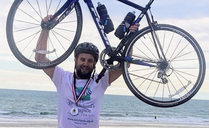Mark Breakwell holds his bicycle above his head. In the background is the sea. He is wearing a St Richard's Hospice branded t-shirt, a cycling helmet, and medal.