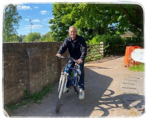 Pete Breakwell sits on his bicycle on a sunny day. The sky is blue, and there are green trees behind him.