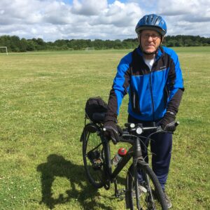 A man stands in a green, grassy field with a black bicycle. He is wearing a blue cycling jacket, black trousers and a blue cycle helmet.