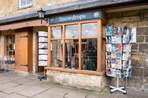 The St Richard's Hospice shop in Broadway. The shop is made from Cotswold stone, and has a blue sign. There is a rack of greeting cards displayed outside.