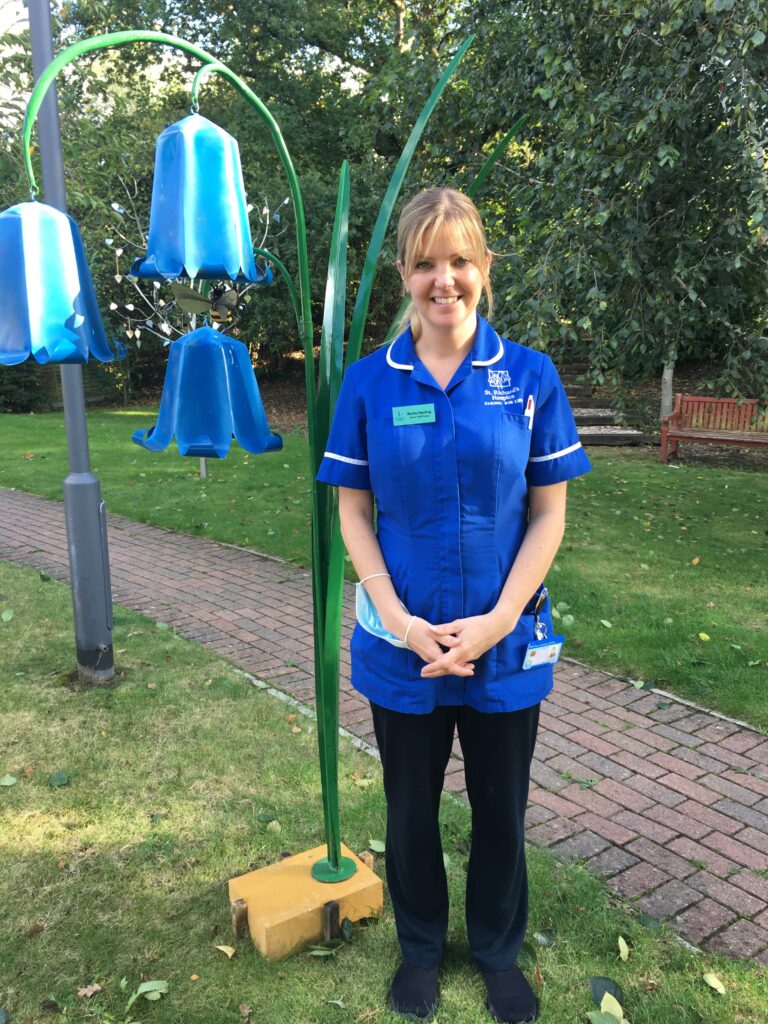 A woman wearing a royal blue nurses' uniform stands in the gardens of St Richard's Hospice. Next to her is a large, metal sculpture of bluebells. She is smiling.