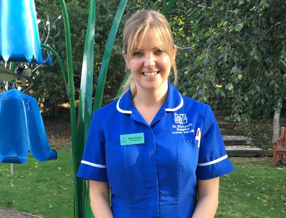 A woman wearing a royal blue nurses' uniform stands in the gardens of St Richard's Hospice. Next to her is a large, metal sculpture of bluebells. She is smiling.