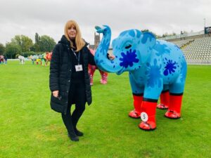 Tricia Cavell, Fundraising Director at St Richard's Hospice, stands with a large blue elephant sculpture on a grassy pitch. She wears a long black coat, black trousers and jumper. She is not smiling. The sky is grey.