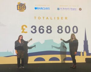 Two women, wearing all black, stand together holding champagne flutes in a toast. They stand in front of a large screen, which has a message reading Totaliser £368,800.