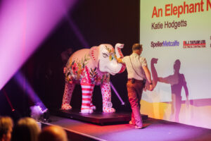 A large pink and red painted elephant sculpture is wheeled on stage by two people wearing beige zoo-keepers clothing. The lighting is bright pink and purple. 
