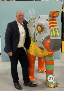 Dale Parmenter wears a dark suit and white shirt and stands next to a large multi-coloured elephant sculpture. He is smiling. 