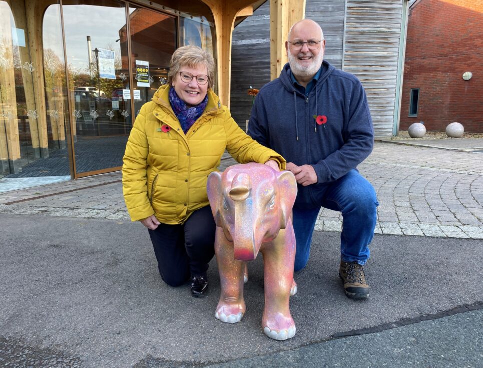 Two people, one wearing black trousers and a yellow coat, the other in jeans and a navy blue jumper, stand together outside St Richard's Hospice. In front of them is a small pink elephant sculpture.