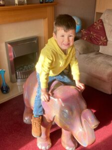 A young boy wearing jeans and a light yellow hoodie sits on a small pink elephant sculpture. He is smiling.