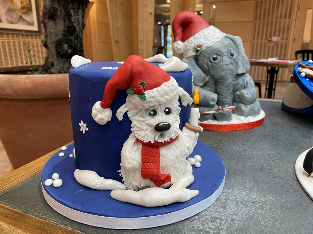 An iced, blue cake featuring a white dog holding a candle and wearing a red Santa hat.