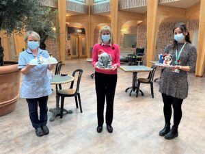 Three people stand together, socially distanced, holding elaborately iced Christmas cakes. They all wear face masks.