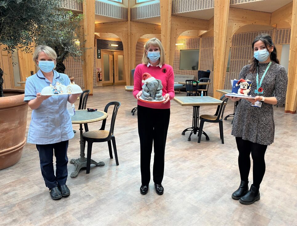 Three people stand together, socially distanced, holding elaborately iced Christmas cakes. They all wear face masks.