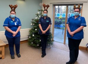 Three hospice nurses wearing black trousers and bright blue uniform tops stand together in the St Richard's Hospice In-patient Unit. Behind them is a Christmas tree, and they all wear reindeer antlers.