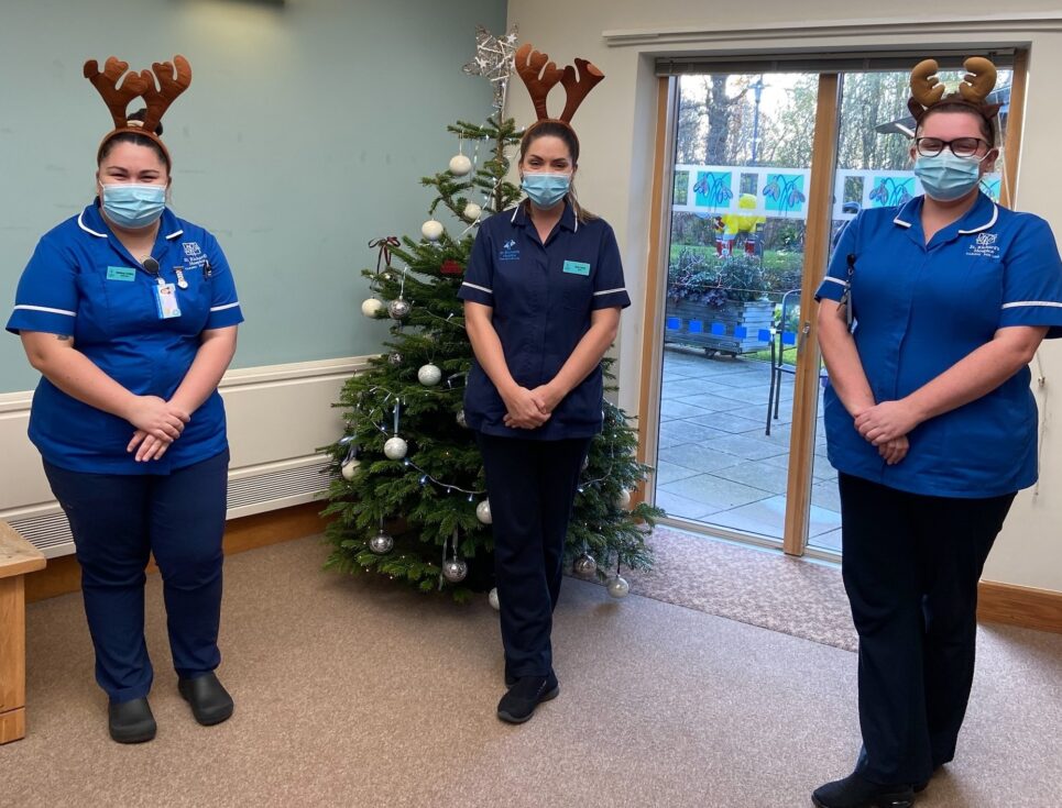 Three hospice nurses wearing black trousers and bright blue uniform tops stand together in the St Richard's Hospice In-patient Unit. Behind them is a Christmas tree, and they all wear reindeer antlers.