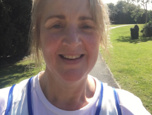 A woman wearing a white t-shirt and blue green running vest smiles as she walks along a path on a sunny day. The grass is green behind her.