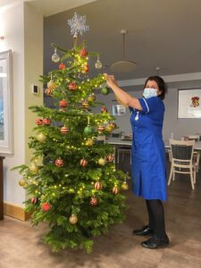 A hospice nurse wearing a blue uniform and face mask puts baubles onto a Christmas tree.