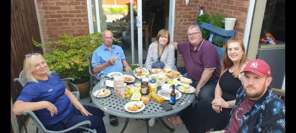 A family sit together round a table in a garden. There is food and drink on the table.