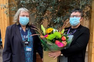 Marina Dixon, wearing a navy blazer and bright blue shirt, is presented with a bunch of pink, yellow, and green flowers by Julie Reece, who wears black. Both Marina and Julie wear face masks.