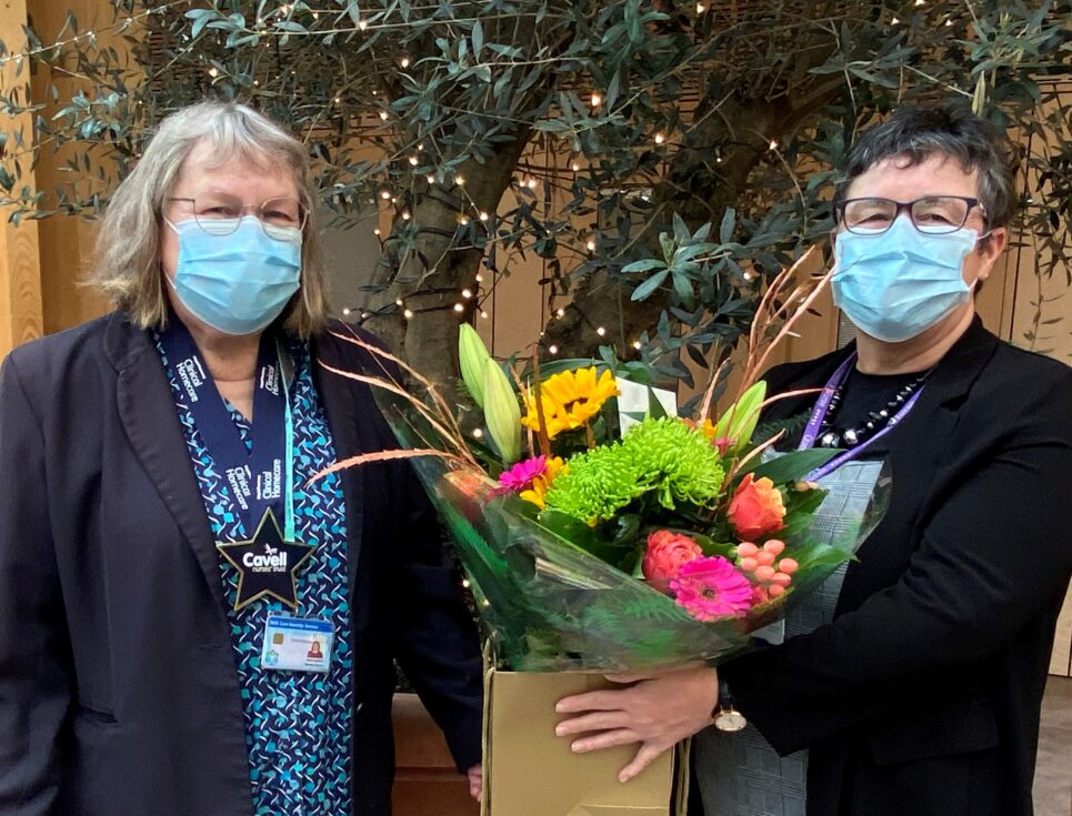 Marina Dixon, wearing a navy blazer and bright blue shirt, is presented with a bunch of pink, yellow, and green flowers by Julie Reece, who wears black. Both Marina and Julie wear face masks.