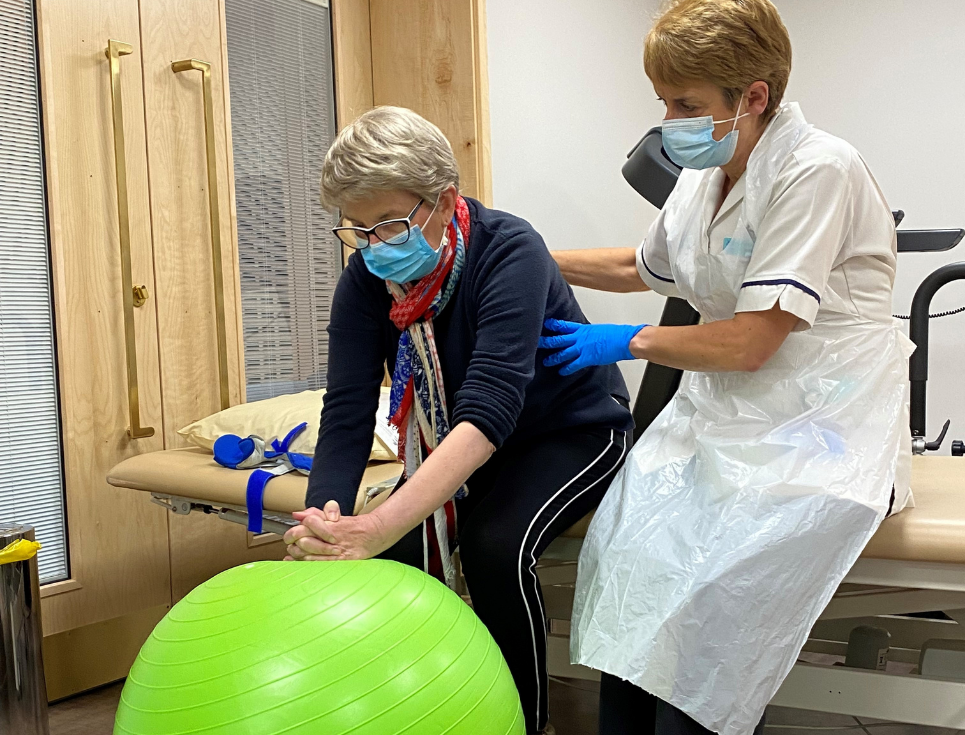 A woman wearing navy clothing and a blue face mask sits and leans forward on a large, green exercise ball. A physiotherapist in a white uniform and blue face mask sits beside her, supporting her.