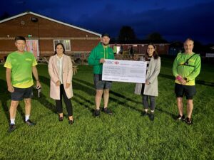 Three people in bright green running clothing present a large cheque to two members of the St Richard's Hospice Fundraising Team. The photo is taken at night, and the sky is dark behind them.