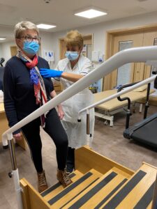 A woman wearing navy clothing and a blue face mask takes part in a physiotherapy session at St Richard's Hospice. A physiotherapist in a white uniform and blue face mask stands beside her, supporting her. 