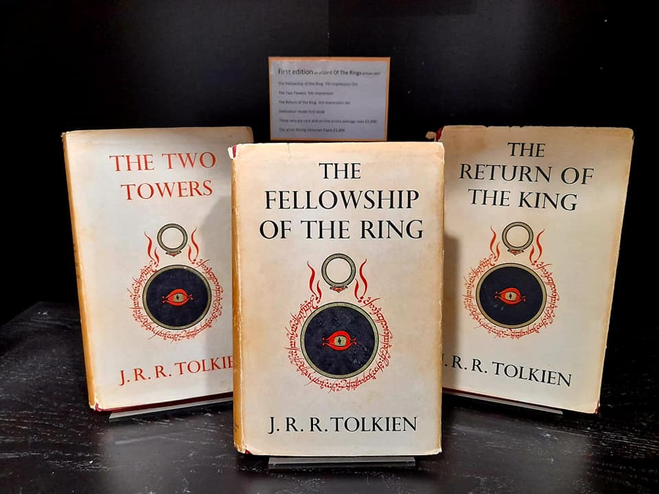 A set of The Lord of the Rings hardback books. Each book is stood upright against a black background. The book covers are pale yellow-white and feature an image of a red eye within a black circle. The text is black and red.