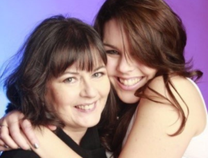 Tricia Bodenham is hugged by her daughter Alice James. Behind them is a purple and pink background. 