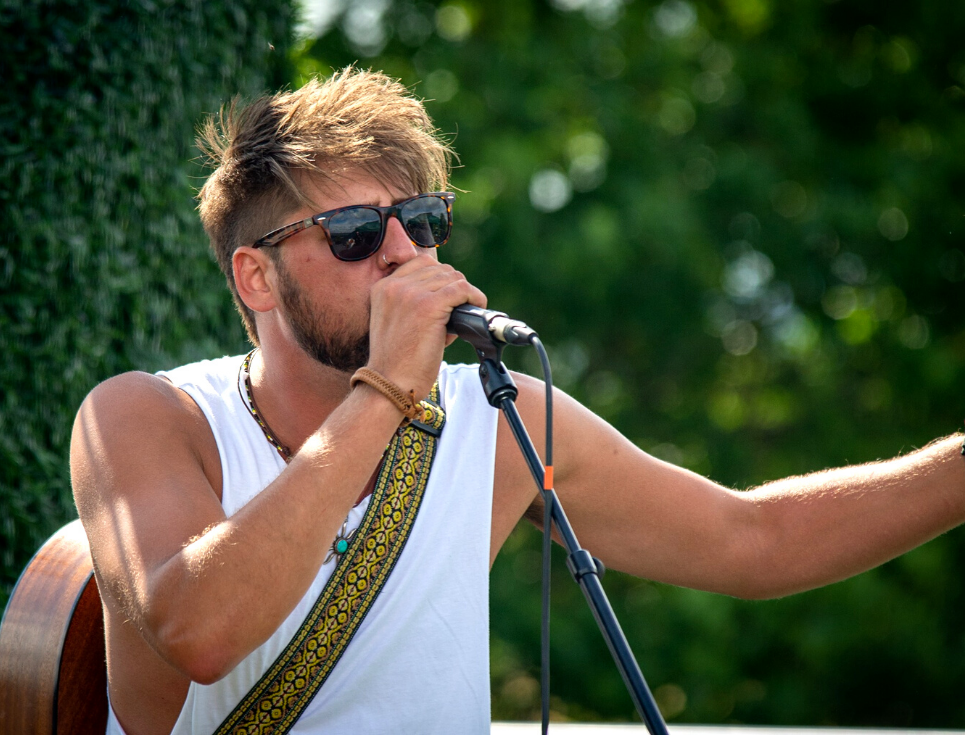 A musician wearing a white vest and black sunglasses sings into a microphone with an acoustic guitar slung across their back.