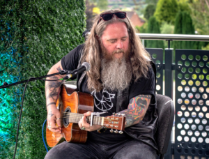 A musician wearing a black t-shirt and black sunglasses on their head sits on a chair, playing an acoustic guitar.