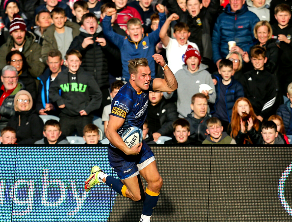A rugby player in a dark blue kit runs holding a rugby ball with a cheering crowd behind them.