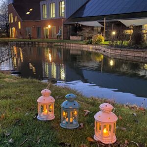 Three small lanterns sit on the grass beside the lake in the gardens at St Richard's Hospice. Each lantern contains a glowing LED light. The photo is taken at dusk.