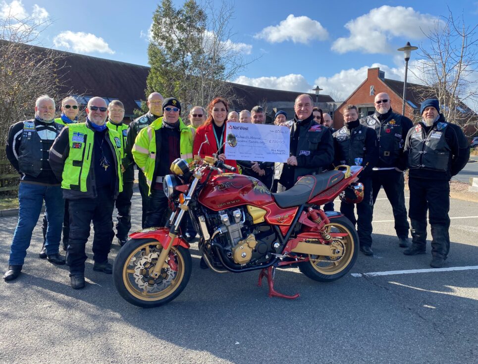 A group of bikers stand together around a red and gold painted motorbike in the car park at St Richard's Hospice. The group present a cheque to a member of the hospice's fundraising team. The sky is blue, with white clouds.