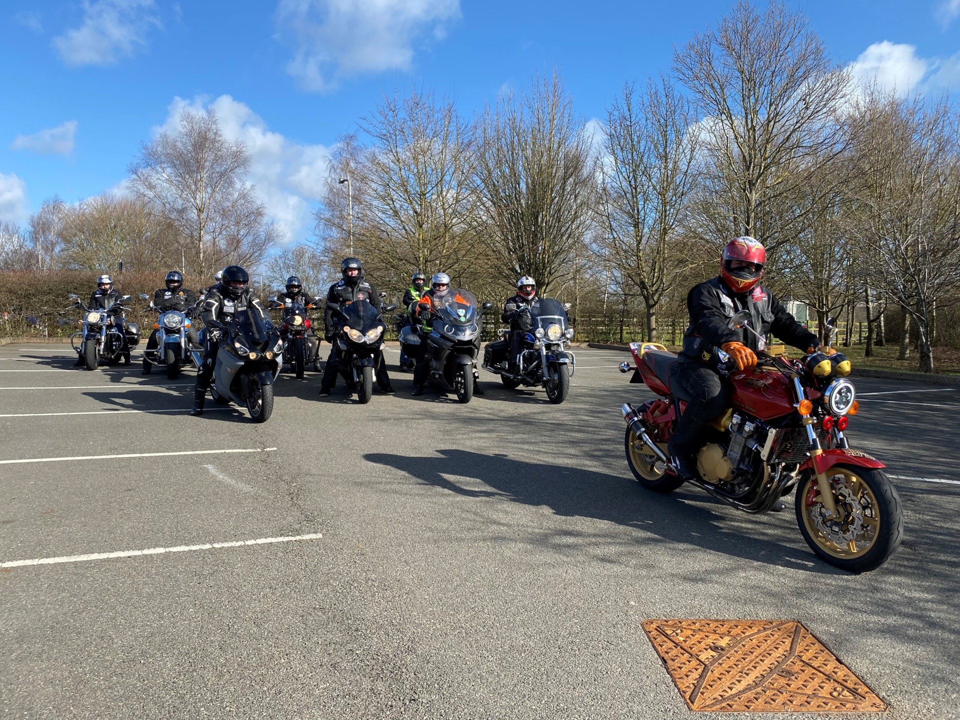 A group of bikers sit on their motorbikes in the car park of St Richard's Hospice. It is a bright and sunny day, with blue skies and white clouds.
