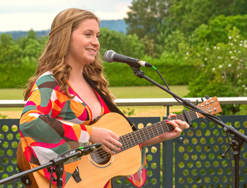 A person wearing a multi-coloured dress stands in front of a microphone playing an acoustic guitar.